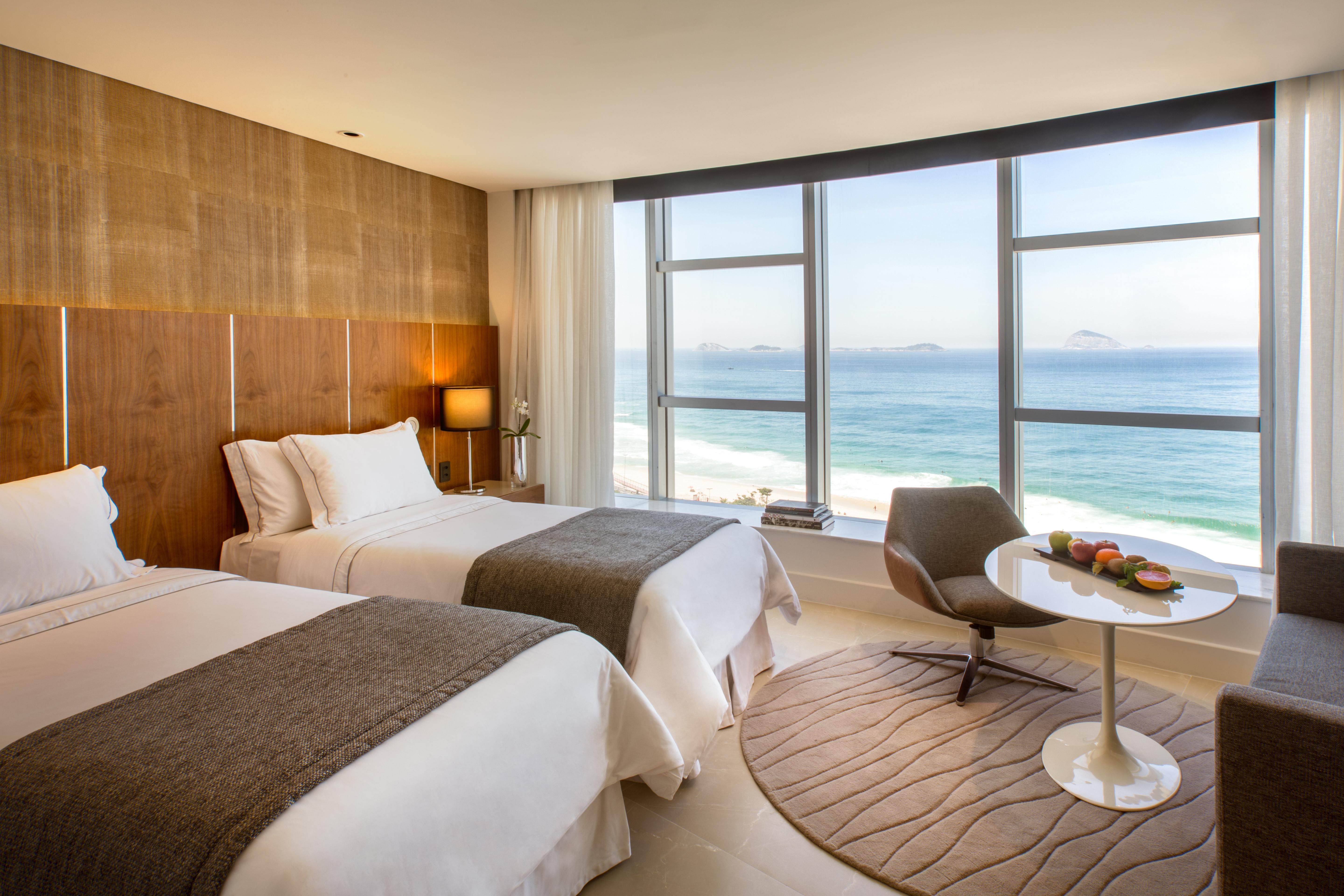 Hotel Nacional Rio De Janeiro - Oficial Exterior photo The photo shows a modern hotel room with two beds adorned in white linens and gray throws. Large windows allow natural light to flood the space, offering a stunning view of the ocean. In the corner, there's a comfortable chair and a small round table